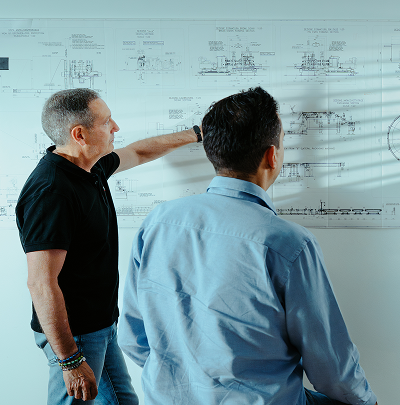 Two engineers reviewing technical machine blueprints on a wall.